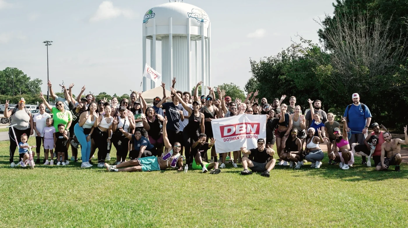 A large, diverse group of Dothan By Nature run club members gathered on a sunny lawn, arms in the air behind a DBN banner