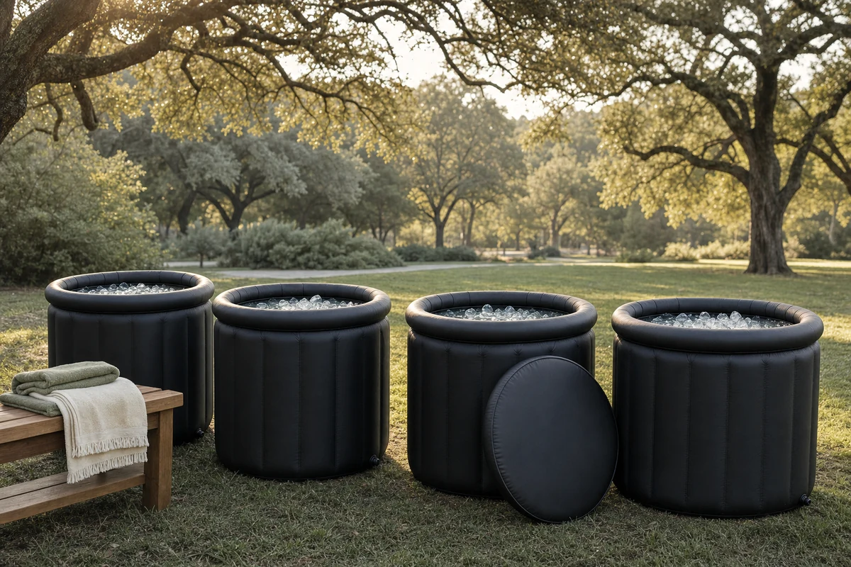 Four portable cold plunge tubs filled with ice water on the grass at a shaded park, with folded towels on a bench nearby